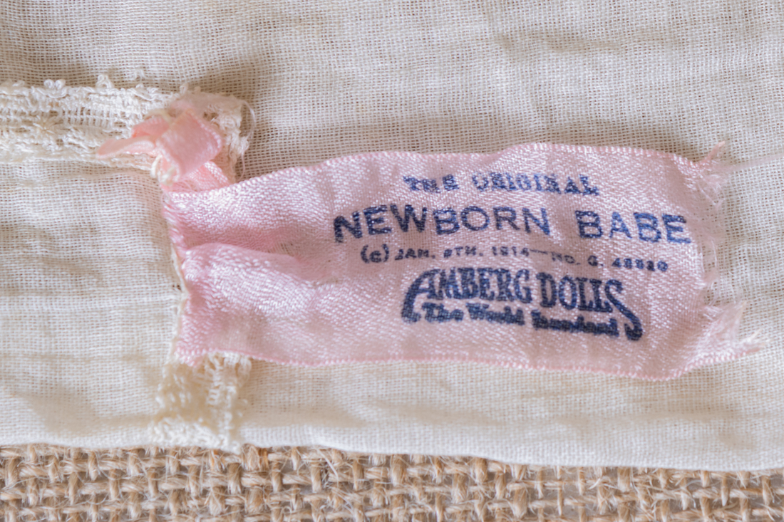 Pink tag on antique baby doll dress marked "the Original Newborn Babe" with the trademark of "Amberg Dolls"